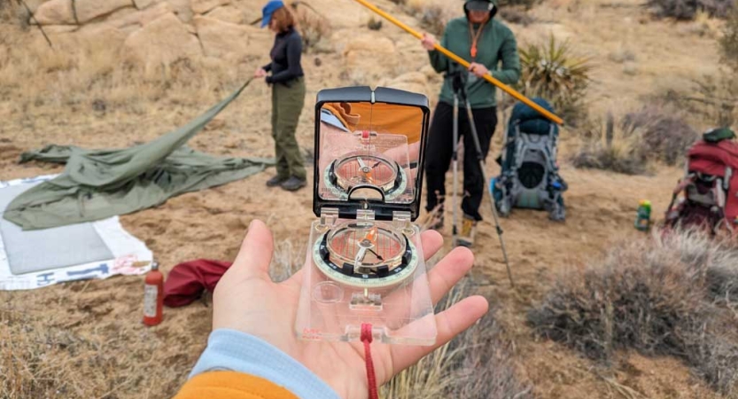 A person holds a compass in the palm of their hand. Behind the hand, there are people working with camp gear. 